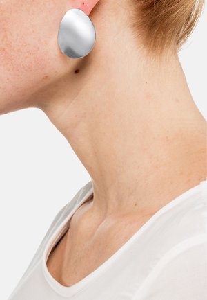 Boucles d'oreilles métalliques argentées avec une surface lisse et réfléchissante dans une forme de goutte d'eau unique. Portées sur un cou à la peau claire avec un haut blanc.