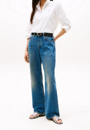 Camicia bianca a maniche lunghe, jeans a gamba larga blu, cintura nera e sandali argento. Presenta un logo sulla camicia, texture in denim e vestibilità rilassata.