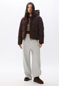 Brązowa kurtka puchowa z kapturem, z grubym wypełnieniem i ściągaczami. W połączeniu z jasnoszarymi szerokimi spodniami dresowymi i ciemnobrązowymi butami.