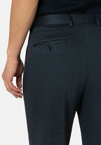 Donkerblauwe, gestructureerde broek met een smalle tailleband, voorzien van een achterzak met een knoop. Het materiaal lijkt geweven en glad.