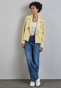 Giubbotto di corduroy giallo con due tasche frontali, abbinato a una maglietta bianca e jeans a gamba larga blu, completato da sneakers bianche.