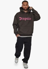 Hoodie noir avec "Dropsize" imprimé en rose, poche kangourou, porté avec un pantalon cargo noir et des baskets grises avec des accents blancs.
