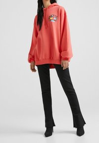 Sudadera oversized de color coral con el texto "PEACE OUT" y un gráfico de sonrisa, combinada con pantalones acampanados negros con dobladillos deshilachados y tacones altos negros.