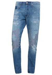 Jeans de mezclilla azul claro con corte slim, que presentan parches desgastados y un ligero desvanecimiento, además de acentos de tela grandes y notables en la parte frontal.