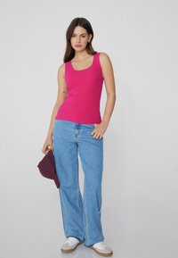 Top sin mangas rosa ajustado hecho de tela elástica, combinado con jeans de pierna ancha azules y zapatillas blancas. Sosteniendo una gorra de béisbol burdeos.