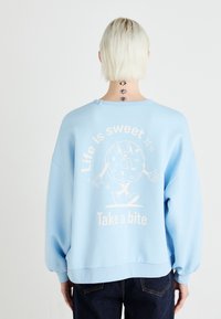 Sweatshirt azul claro com um corte oversized, apresentando uma gráfica divertida de donut e o texto "A vida é doce, dê uma mordida" nas costas.