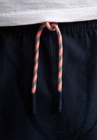 Shorts de marine avec un cordon tressé multicolore comportant des accents noirs et orange ainsi que des embouts en plastique. Coutures visibles à la ceinture.