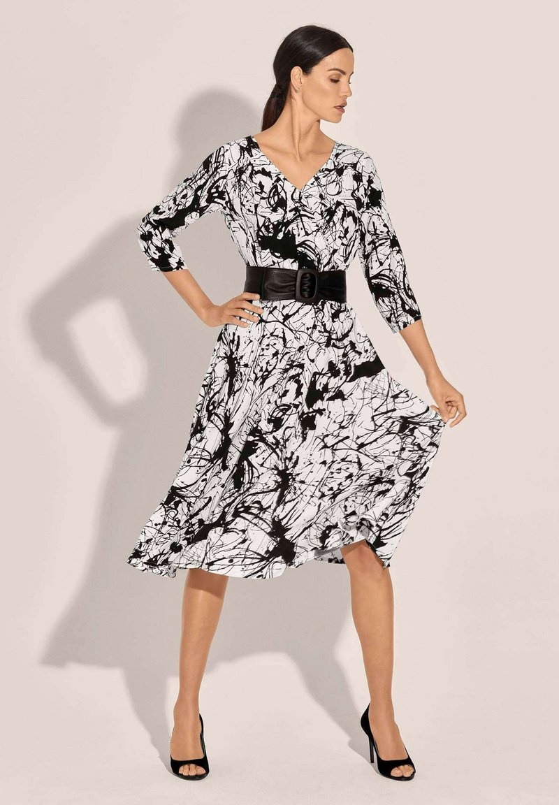Schwarz-weißes Kleid mit drei Viertelärmeln, V-Ausschnitt, abstraktem Spritzer-Muster, tailliert mit einem breiten schwarzen Gürtel, schwingendem Rock, getragen mit schwarzen High Heels.