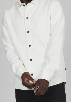 Homme portant une chemise blanche à manches longues boutonnée, ajustant le poignet avec les deux mains, visible des épaules aux hanches sur fond uni.
