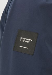 Tejido azul marino con una etiqueta rectangular negra que muestra las coordenadas "55° 40' NORTE 12° 36' ESTE" y "JACK&JONES©1990" en texto blanco.