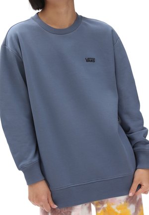 Personne portant un sweat-shirt Vans bleu avec poignets et ourlet côtelés, associé à un pantalon coloré tie-dye, coupé au niveau du cou et des genoux.
