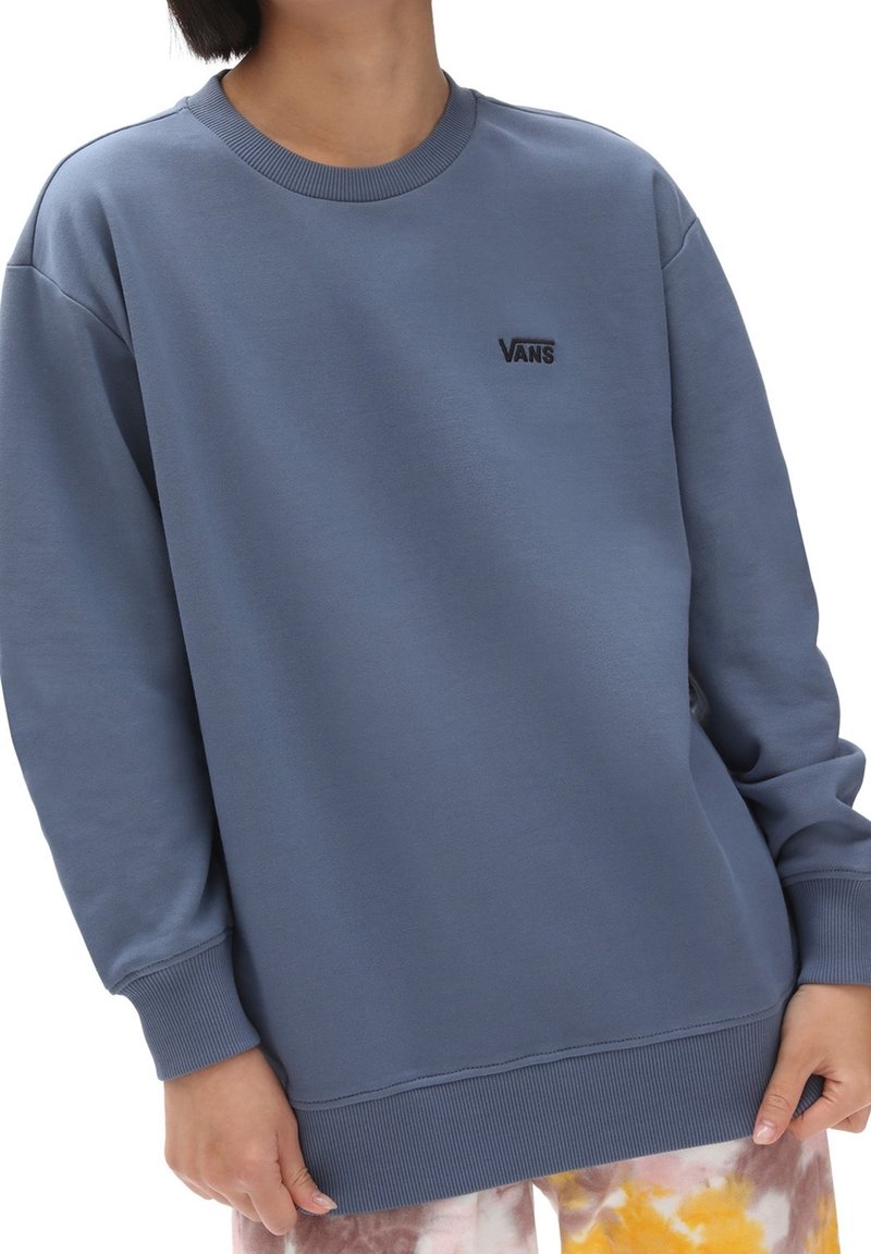 Personne portant un sweat-shirt Vans bleu avec poignets et ourlet côtelés, associé à un pantalon coloré tie-dye, coupé au niveau du cou et des genoux.