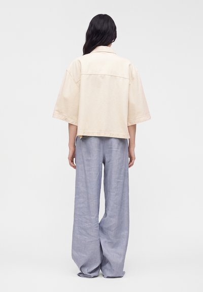 Femme aux longs cheveux foncés debout, portant une veste ample crème à manches courtes et un pantalon large gris clair, de dos sur un fond uni.