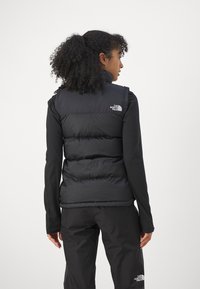 Μαύρο αμάνικο μπουφάν με ψηλό γιακά, διαθέτοντας σχεδιασμό με ραφές και λογότυπο στον αριστερό ώμο, φορεμένο πάνω από ένα μακρυμάνικο τοπ.