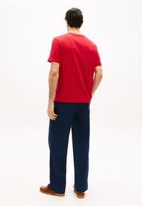 Άνδρας που στέκεται με την πλά τη γυρισμένη, φοράει κόκκινο κοντομάνικο πουκάμισο, σκούρο μπλε παντελόνι και καφέ παπούτσια σε λευκό φόντο.