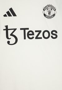 Maillot de football blanc avec logo Adidas noir, texte "Tezos" et blason de Manchester United en haut à droite. Tissu lisse avec un design simple.