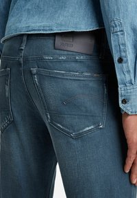 Tmavě modré džíny z denimu v úzkém střihu, s jemným vyblednutím a černou koženou nášivkou na pase s názvem značky "RAW."