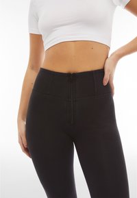 Nainen pukeutuneena mustiin korkeavyötäröisiin leggingsseihin, joissa on etuvetoketju, ja valkoiseen lyhyeen toppiin, käsi lanteilla, valkoista taustaa vasten.