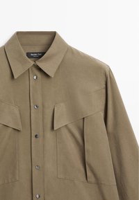 Khaki button-up shirt with a classic collar, two large chest pockets, and black snap buttons. Textured fabric offers a structured look.