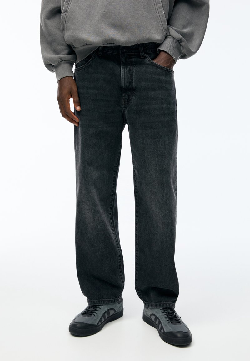 Personne portant un jean noir ample, un sweat-shirt gris et des baskets gris et noir, debout avec une main dans la poche sur fond blanc.