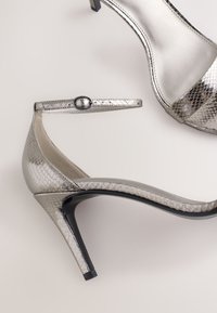 Srebrne sandały na obcasie z fakturą skóry węża, wyposażone w regulowany pasek na kostkę oraz elegancki spiczasty nosek. Czarna podstawa obcasa.