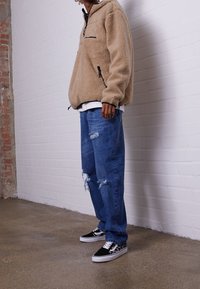 Pullover in pile color sabbia con zip frontale e tasche laterali, abbinato a jeans blu distressed e sneakers nere con accenti bianchi.