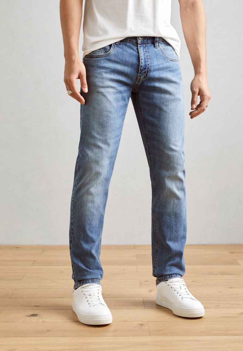 Light blue denim jeans with a straight fit, faded texture, and five-pocket design. Worn with white sneakers on a wooden floor.