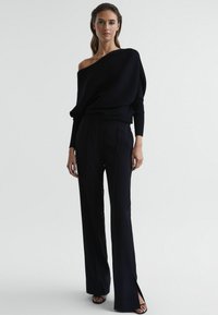Zwarte geribbelde off-shoulder top gecombineerd met hoge taille, op maat gemaakte zwarte broeken met zijsplitten, aangevuld met zwarte hakken.