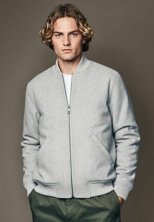 Graue Bomberjacke mit glatter Textur, kontrastierendem geripptem Kragen und Bündchen, Frontreißverschluss und Seitentaschen. Kombiniert mit olivgrünen Hosen.