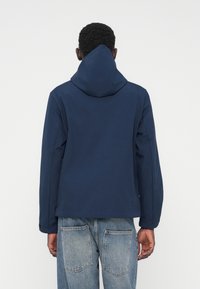 Mørkeblå hættejakke med en glat tekstur, der har et minimalistisk design og kontrasterende syninger. Bæres med lyseblå jeans.