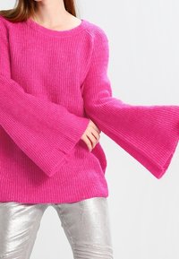 Pull tricot rose vif avec manches larges en forme de cloche. Texture côtelée, encolure arrondie, associé à un pantalon argenté brillant.