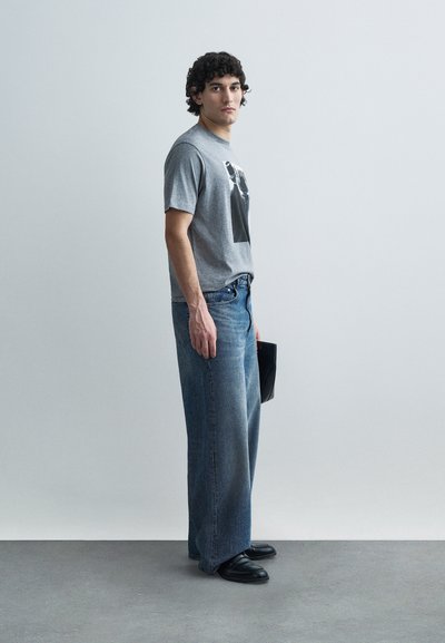 Γκρι βαμβακερό t-shirt με γραφικό σχέδιο, φαρδιά μπλε τζιν, μαύρα δερμάτινα παπούτσια και ένας μαύρος φάκελος στο χέρι, στέκεται μπροστά σε ουδέτερο φόντο.