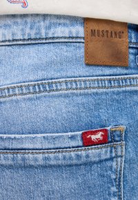 Jasnoniebieskie dżinsy z skórzaną metką MUSTANG z tyłu. Posiadają czerwoną naszywkę z białym emblematem konia na kieszeni.