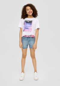 Weißes Grafik-T-Shirt mit buntem Text, kombiniert mit Denim-Shorts und einem pinken Gürtel. Das Model trägt weiße Sneakers und hat lockige Haare.