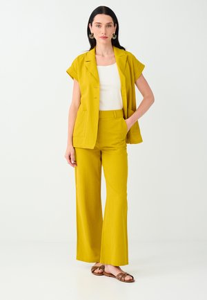 Vrouw met zwart haar draagt een mosterdgele blazer met korte mouwen en broek met wijde pijpen, een wit topje, grote ronde oorbellen en bruine sandalen.
