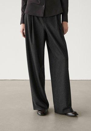 Persoon die donkergrijze wijde broek draagt, gecombineerd met een zwart overhemd met knopen en zwarte leren schoenen, staand op een neutrale vloer.