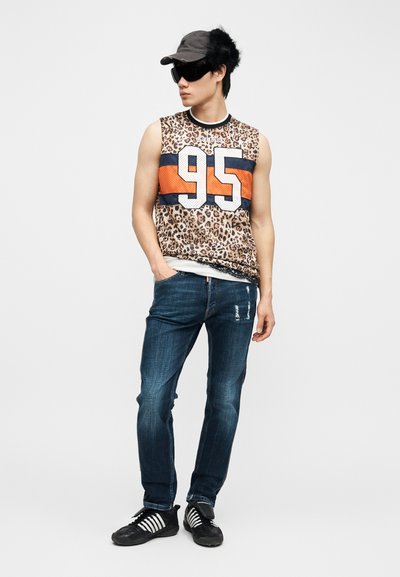 Jeune homme portant un haut sans manches à motif léopard avec le numéro 95, un jean foncé, une casquette noire et des lunettes de soleil, debout avec une main dans la poche.