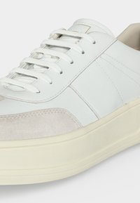 Gros plan d'une basket en cuir blanche avec des lacets blancs et un détail en daim beige sur une semelle épaisse blanc cassé.