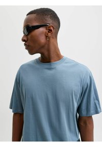 Niebieska bawełniana koszulka z krótkim rękawem i okrągłym dekoltem. Charakteryzuje się gładką fakturą i luźnym krojem, noszona z czarnymi okularami przeciwsłonecznymi.