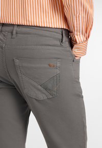 Sive hlače z teksturirano tkanino, ki vključuje edinstven dizajn žepov z kontrastnim delom in okrasnim šivanjem.