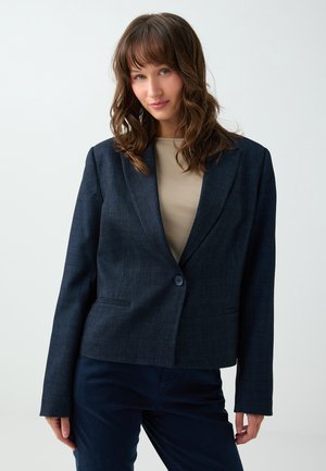 Jeune femme aux cheveux bruns ondulés portant un blazer bleu foncé sur un haut beige, debout devant un fond clair uni.