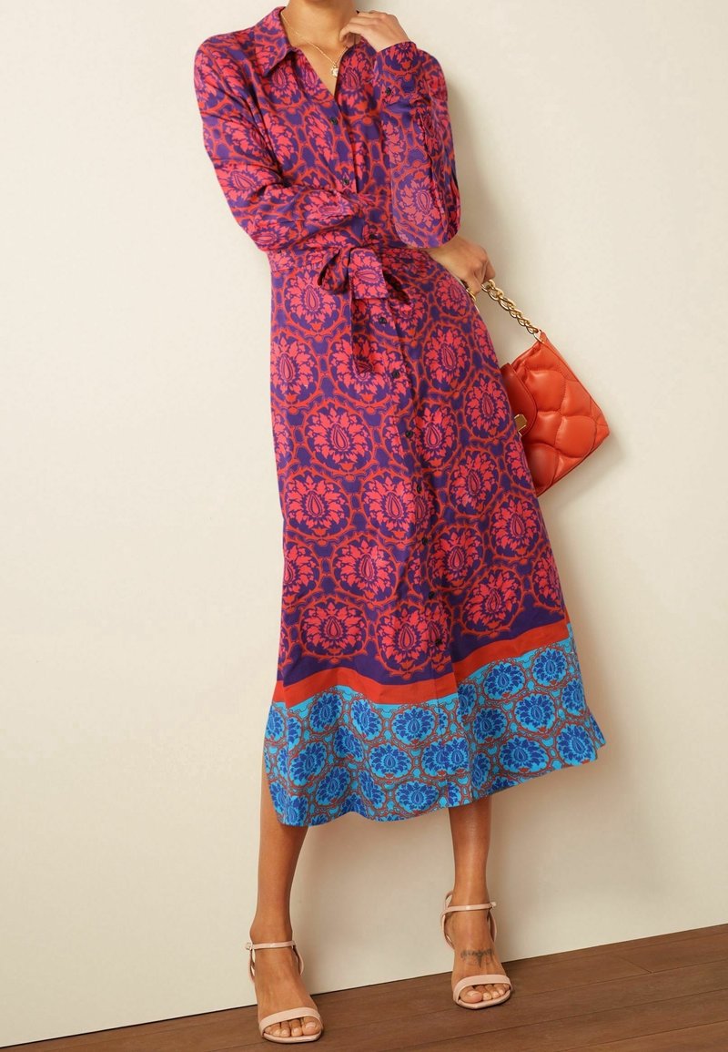 Femme portant une robe violette à manches longues avec des motifs floraux rouges et bleus, tenant une pochette orange, debout sur un sol en bois.