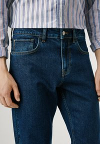 Jeans de mezclilla azul oscuro con un diseño tradicional de cinco bolsillos, cierre de botón de latón y detalles de costura en naranja contrastante.