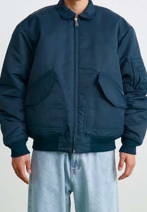 Mand iført mørkeblå polstret bomberjakke med to forlommer med klap og lyseblå jeans, stående mod en ensfarvet baggrund.