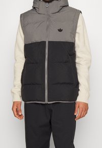 Vadderad väst i grått och svart med huva, framdragkedja och liten svart logotyp. Tillverkad av tyg med en slät yta.