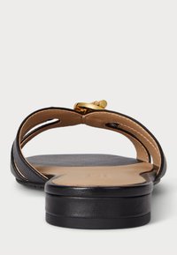 Juodos plokščios šlepetės su minimalistiniu dizainu, turinčios lygaus odos paviršiaus, subtilių siūlių ir auksinės spalvos sagties akcentu šalia kulno.