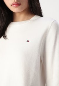 Maglione bianco realizzato in morbido tessuto a maglia, con scollatura tonda e un piccolo logo ricamato in rosso, bianco e blu sul petto.