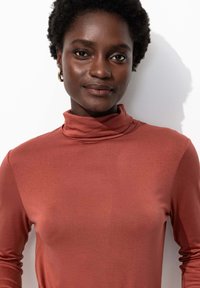 Haut à col roulé à manches longues de couleur rouille, en tissu doux avec une texture lisse, présentant une silhouette ajustée et un décolleté subtil.