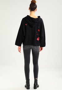 Kvinna med hår i knut som bär en svart huvtröja med röda läppar och stjärnpatchar, grå tajta jeans och svarta ankelboots, står vänd bort.