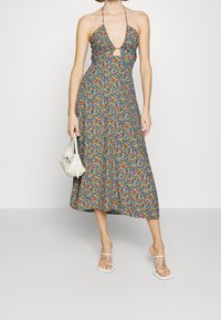 Květinové šaty s halter krkem a výřezem, mnohobarevný potisk na modrém pozadí, splývající midi délka, nošeny s malou bílou taškou.